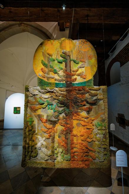 Liela formāta tekstilmākslas darbs ekspozīcijā ar koka un putnu lidojuma motīviem, veidots reljefā tehnikā siltos dzeltenos, zaļos un oranžos toņos.