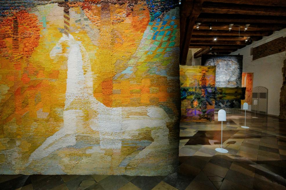 Tekstilmākslas darba fragments ar reljefu auduma struktūru un siltu dzeltenu, oranžu un zemes toņu krāsu laukumiem, izstādīts muzeja vēsturiskajā interjerā.