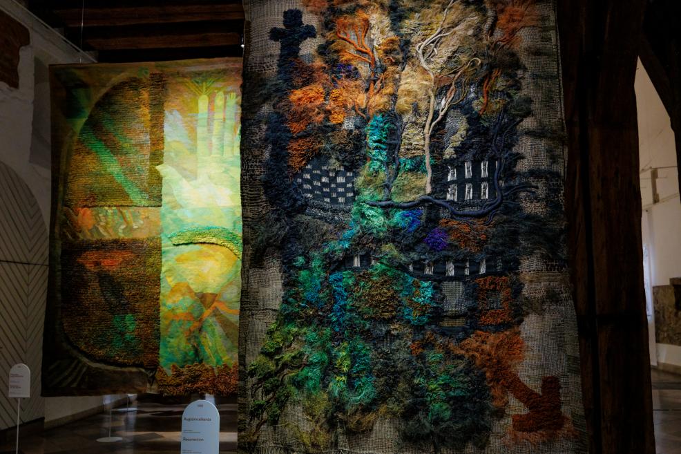 Piekārts tekstilmākslas darbs ar bagātīgu reljefu, kur tumši zilie, zaļie, brūnie un oranžie pavedieni veido krusta un koka sakņu asociācijas muzeja ekspozīcijā.