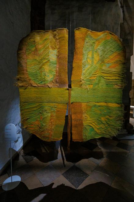 Četrās daļās sadalīts vertikāls tekstilmākslas darbs zaļos, dzeltenos un okera toņos ar izteiktu reljefu, piekārts ar trosēm muzeja telpā pie akmens sienas.