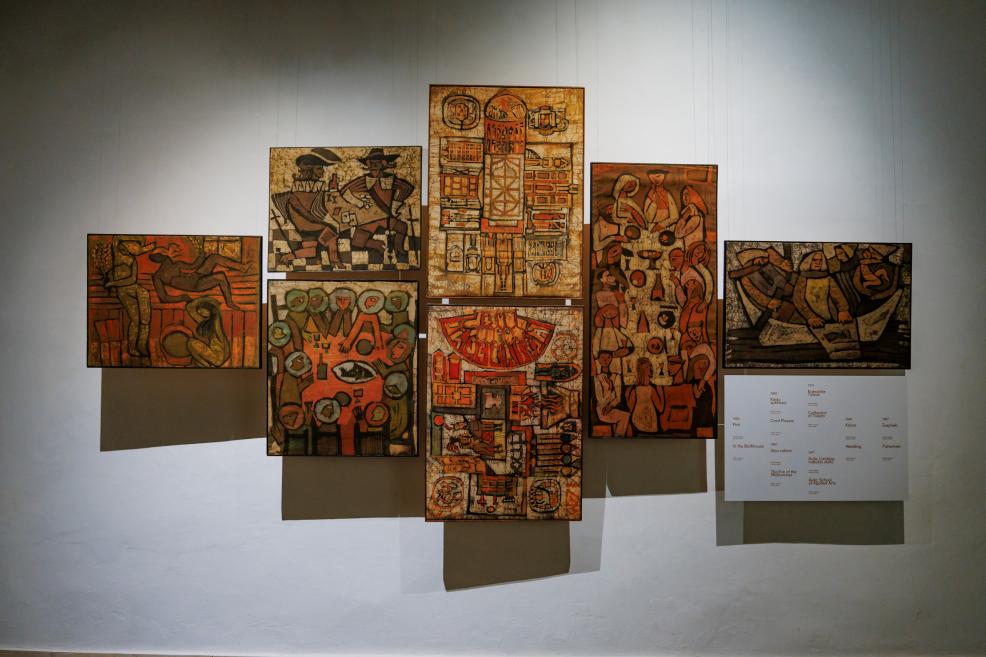 Izstādes ekspozīcijas skats ar vairākiem sienā eksponētiem darbiem siltā, vāji apgaismotā telpā.