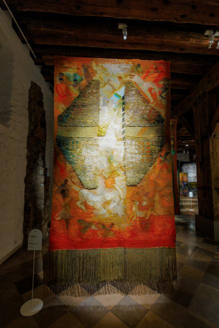 Liela formāta tekstilmākslas darbs ar reljefu virsmu un siltu oranžu, dzeltenu un zemes toņu kolorītu, eksponēts vēsturiskā telpā ar koka sijām.