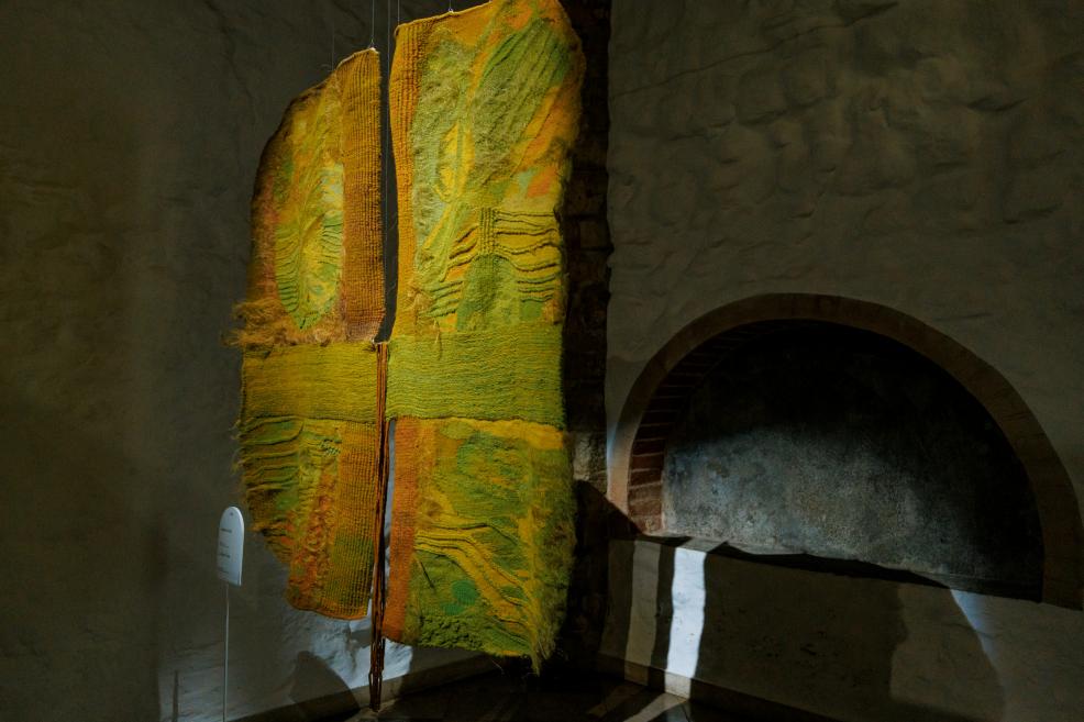 Telpā pie akmens sienas eksponēts vertikāls tekstilmākslas darbs zaļi dzeltenos toņos ar reljefainu faktūru, sadalīts četrās daļās un piekārts ar trosēm.