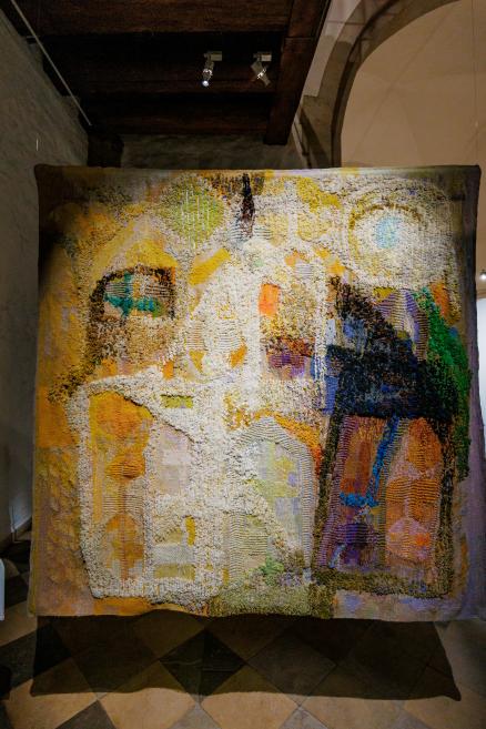 Liels piekārts tekstilmākslas darbs ar bagātīgu reljefu, kurā krēmkrāsas, dzelteni, zaļi un zilgani laukumi veido abstraktu, mozaīkai līdzīgu kompozīciju muzeja telpā.