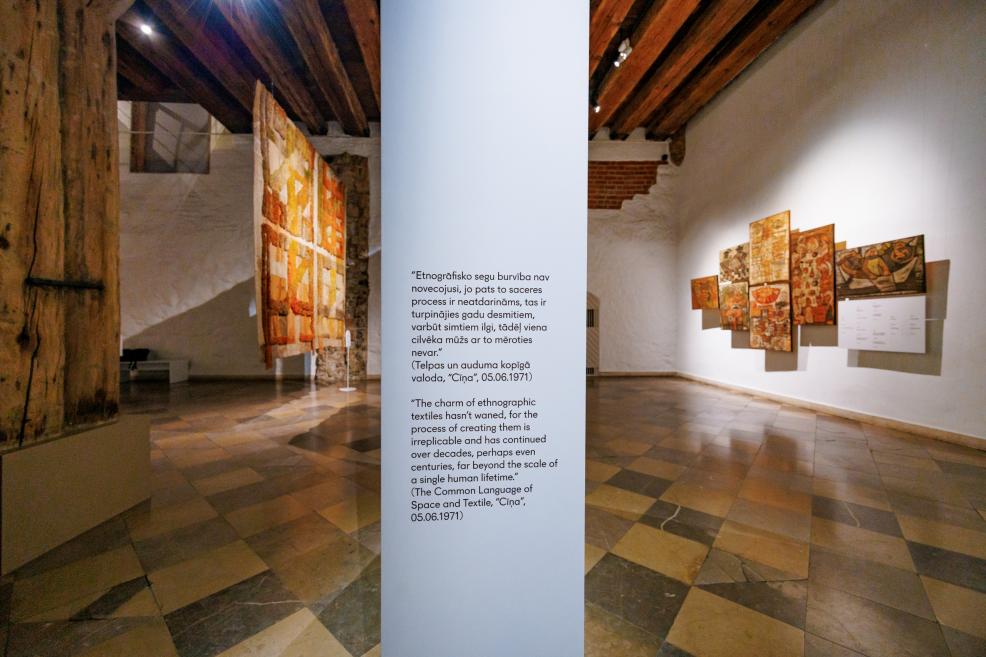 Muzeja ekspozīcijas skats ar piekārtu tekstilmākslas darbu, sienā izvietotu darbu grupu un vertikālu sienas paneli ar latviešu un angļu valodā lasāmu citātu.