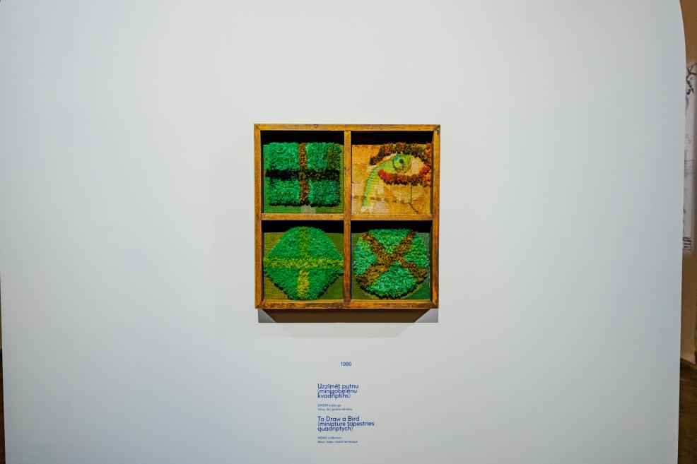 Tekstilmākslas darbs četrdaļīgā koka rāmī ar zaļos un dzeltenos toņos veidotiem simboliskiem motīviem, eksponēts uz baltas sienas ar informatīvu uzrakstu zem tā.