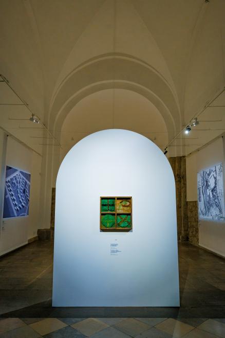 Ekspozīcijas skats ar nelielu tekstilmākslas objektu četrdaļīgā koka rāmī, kas eksponēts nišveida arhitektoniskā sienā muzeja zālē ar velvēm.