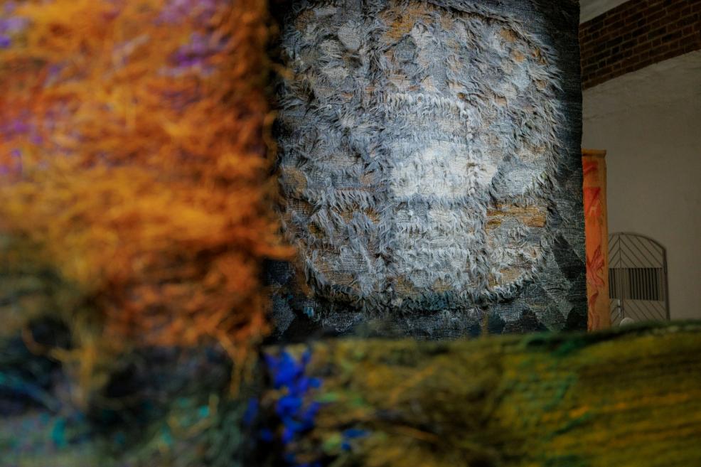 Tekstilmākslas darba fragments ekspozīcijā, kur priekšplānā redzamas krāsainas, fakturētas šķiedras, bet fonā – liela, pelēcīga tekstilija ar reljefu virsmu.