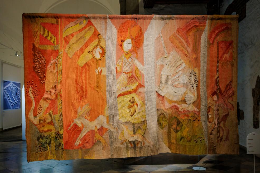 Liela formāta tekstilmākslas darbs ekspozīcijā ar figurāliem motīviem siltos oranžos, dzeltenos un bēšos toņos, kur redzami stilizēti tēli un bagātīga reljefa faktūra.