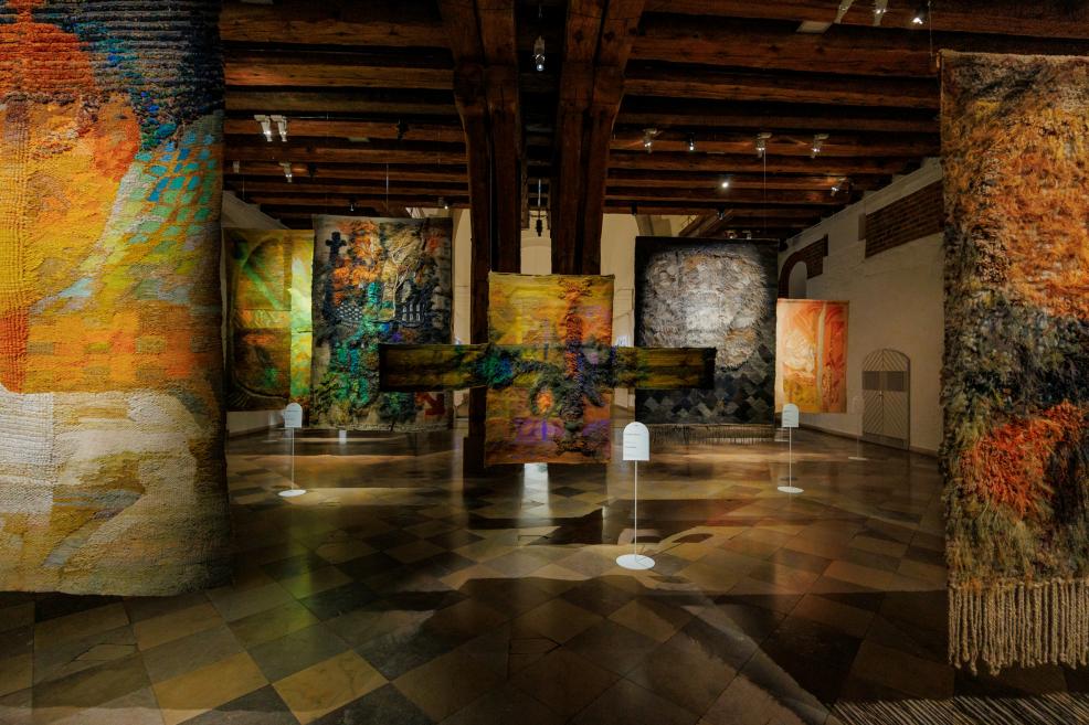 Tekstilmākslas izstādes ekspozīcijas kopskats ar vairākiem monumentāliem, telpiski izvietotiem tekstiliju darbiem muzeja zālē ar koka sijām un vēsturisku grīdas segumu.