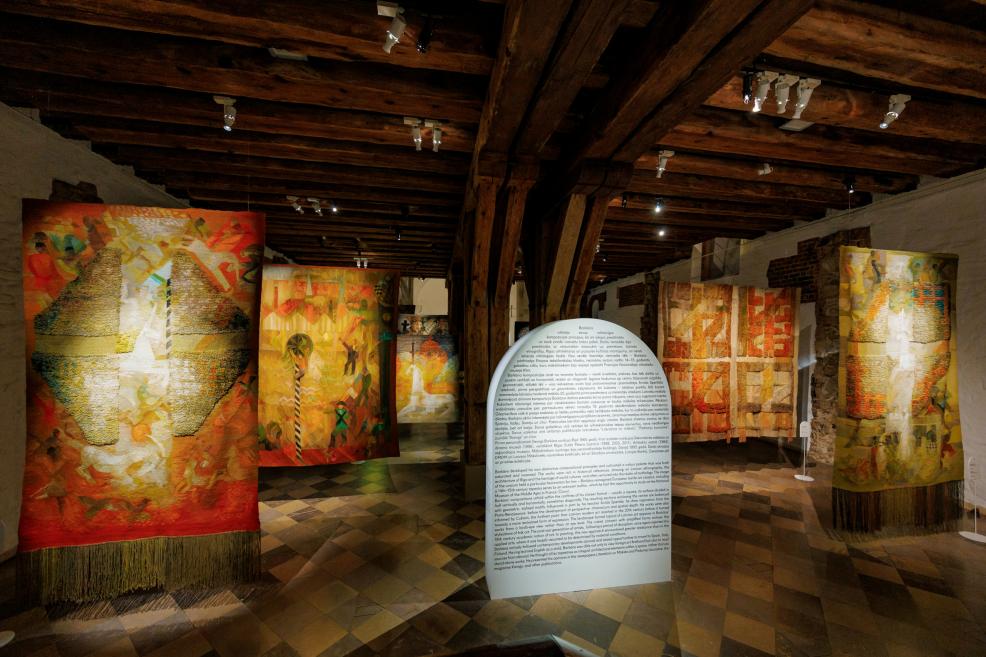 Tekstilmākslas darbu ekspozīcija vēsturiskā telpā ar koka sijām un akmens grīdu.