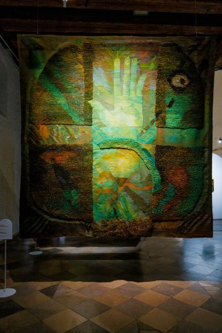 Liels piekārts tekstilmākslas darbs ar izteiktu reljefu, kur zaļgani, zilgani un okera toņi veido krusta formu un stilizētu rokas motīvu muzeja ekspozīcijā.