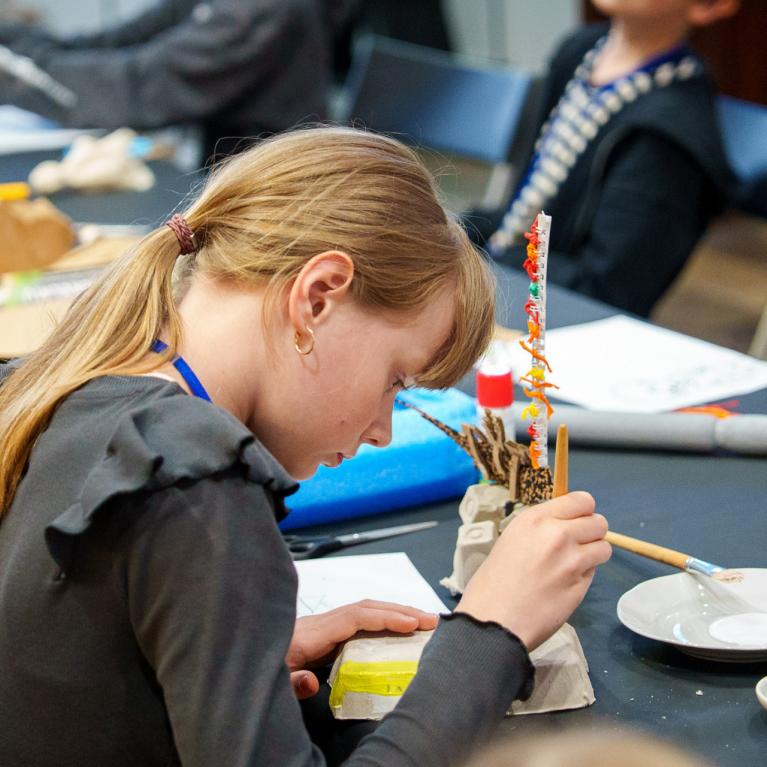 Meitene ar zirgasti un pelēku džemperi piedalās radošā darbnīcā, gleznojot ar otu pie galda ar krāsām, traukiem un dažādiem amatniecības materiāliem.