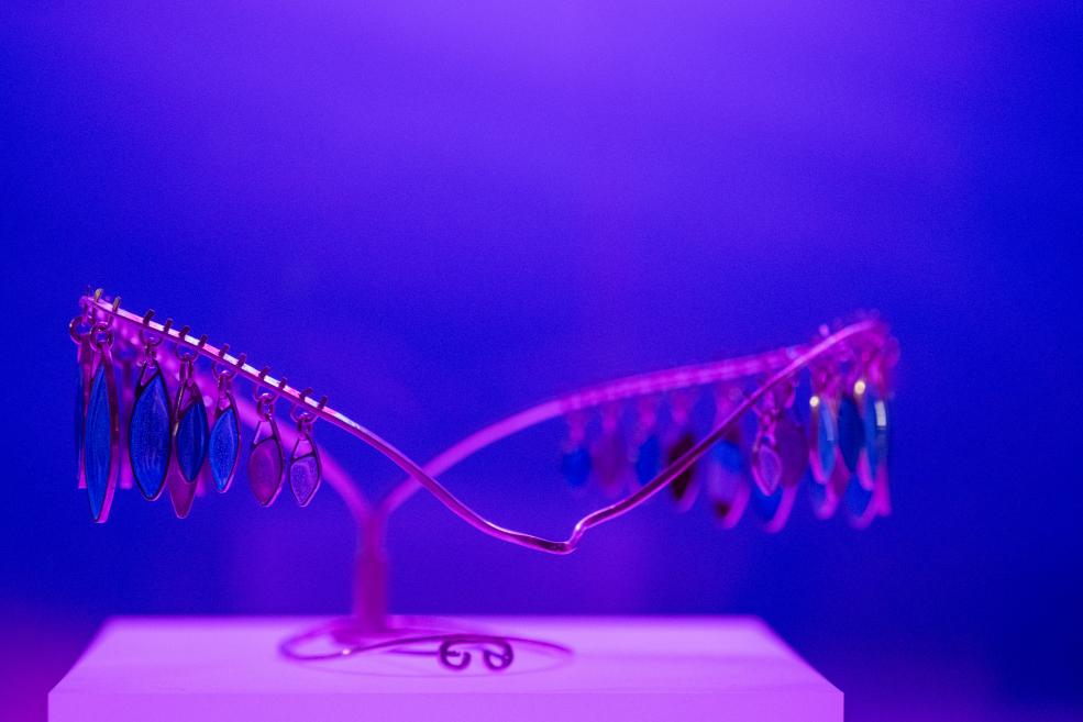 A pink metal structure with hanging blue and pink oval elements on a white pedestal against a violet-blue background.