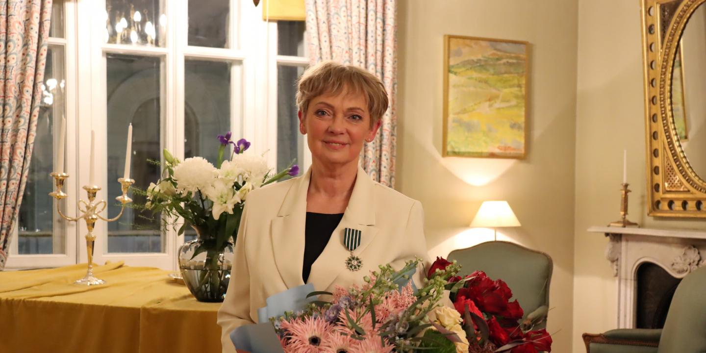 A woman wearing a light-coloured jacket with an order insignia and holding a bouquet stands in a classical interior with candles and flowers.