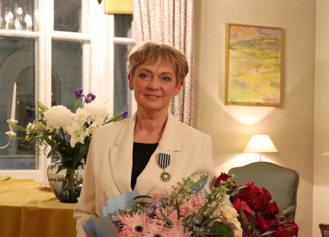 A woman wearing a light-coloured jacket with an order insignia and holding a bouquet stands in a classical interior with candles and flowers.