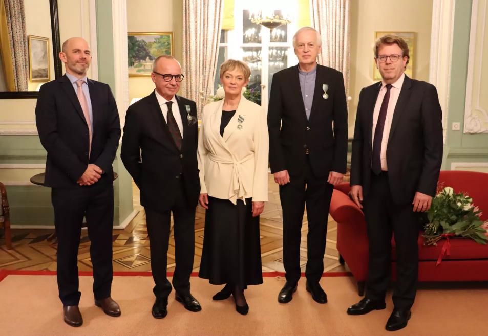 Grupas portrets formālā pasākumā reprezentatīvā interjerā, kur piecas personas pozē kamerai.