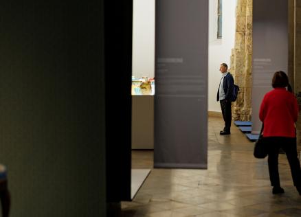 Muzeja zālē apmeklētāji apskata ekspozīciju; priekšplānā daļēji redzams dekoratīvs krēsls, fonā vitrīna ar stikla traukiem un uz pjedestāla novietots stikla galdiņš.