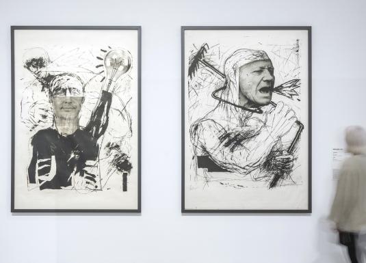 Baltā izstāžu zālē pie sienas eksponēti divi liela formāta melnbalti grafikas darbi ar ekspresīvām cilvēka figūrām, kuru priekšā kustībā izplūdis apmeklētājs aplūko darbu anotāciju.