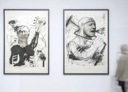 Baltā izstāžu zālē pie sienas eksponēti divi liela formāta melnbalti grafikas darbi ar ekspresīvām cilvēka figūrām, kuru priekšā kustībā izplūdis apmeklētājs aplūko darbu anotāciju.