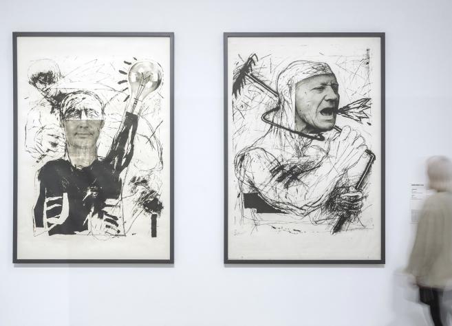 Baltā izstāžu zālē pie sienas eksponēti divi liela formāta melnbalti grafikas darbi ar ekspresīvām cilvēka figūrām, kuru priekšā kustībā izplūdis apmeklētājs aplūko darbu anotāciju.