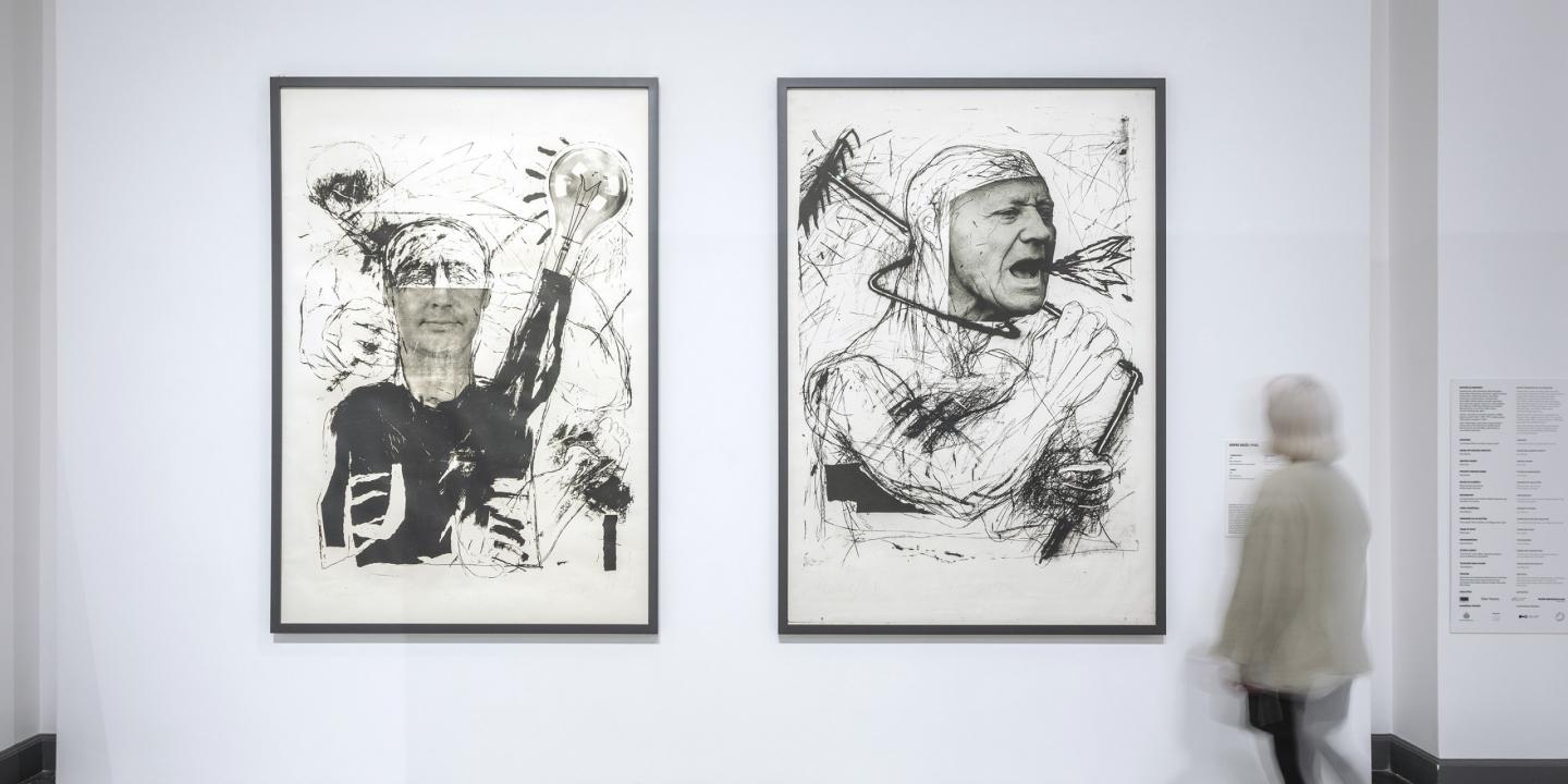 Baltā izstāžu zālē pie sienas eksponēti divi liela formāta melnbalti grafikas darbi ar ekspresīvām cilvēka figūrām, kuru priekšā kustībā izplūdis apmeklētājs aplūko darbu anotāciju.