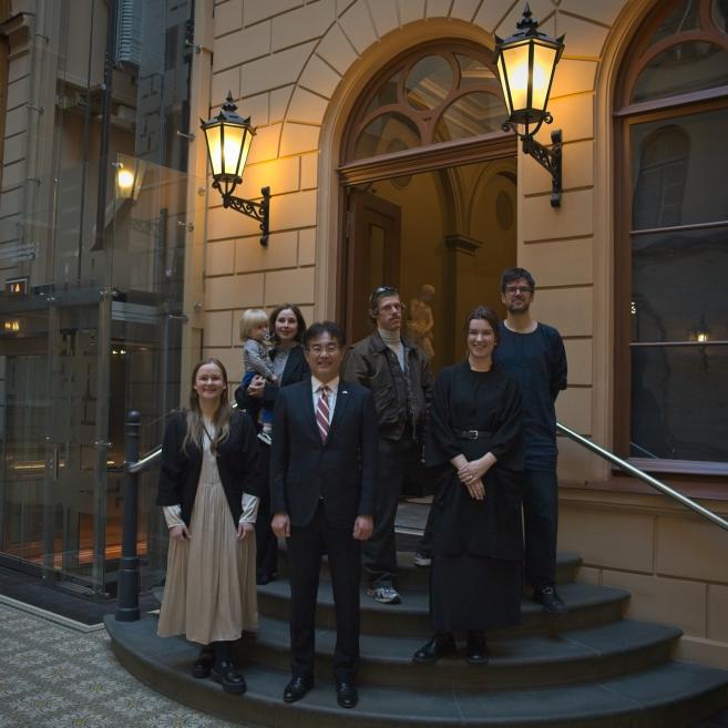 Seši pieaugušie un viens bērns stāv uz kāpnēm pie vēsturiskas ēkas ieejas zem divām iedegtām laternām un pozē kopīgai fotogrāfijai.