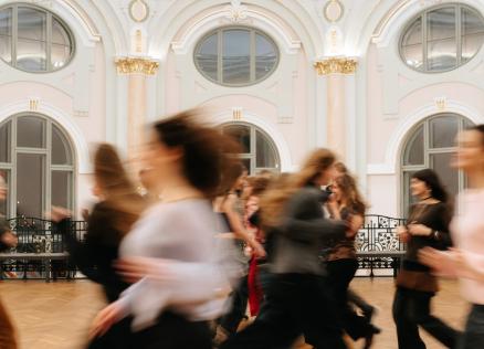 Plašā, greznā zālē ar augstiem arkveida logiem kustībā izplūduši cilvēki skrien vai pārvietojas, radot dinamisku, miglainu efektu.
