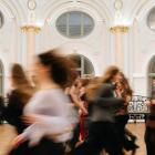 Plašā, greznā zālē ar augstiem arkveida logiem kustībā izplūduši cilvēki skrien vai pārvietojas, radot dinamisku, miglainu efektu.