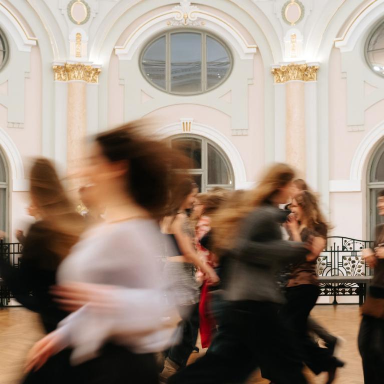 Plašā, greznā zālē ar augstiem arkveida logiem kustībā izplūduši cilvēki skrien vai pārvietojas, radot dinamisku, miglainu efektu.