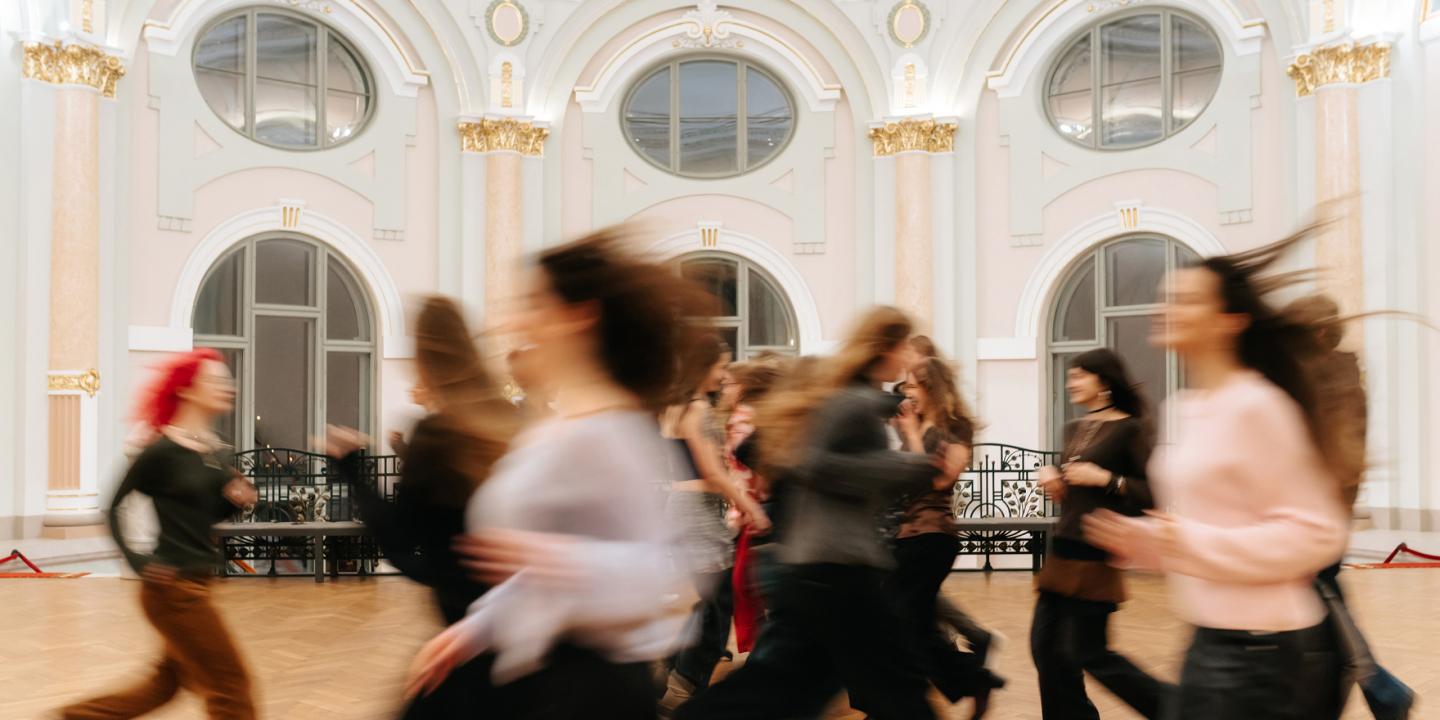 Plašā, greznā zālē ar augstiem arkveida logiem kustībā izplūduši cilvēki skrien vai pārvietojas, radot dinamisku, miglainu efektu.