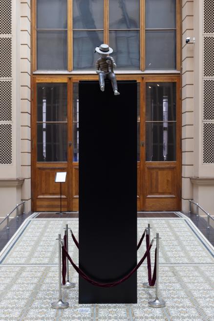 A monochrome doll-like boy figure wearing a straw hat sits on a tall black pedestal in a museum hall.