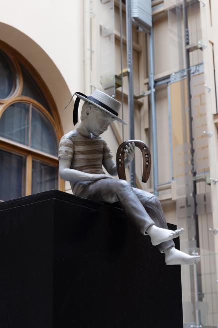An installation with a seated boy sculpture wearing a hat and holding a horseshoe on a black pedestal in a museum interior.