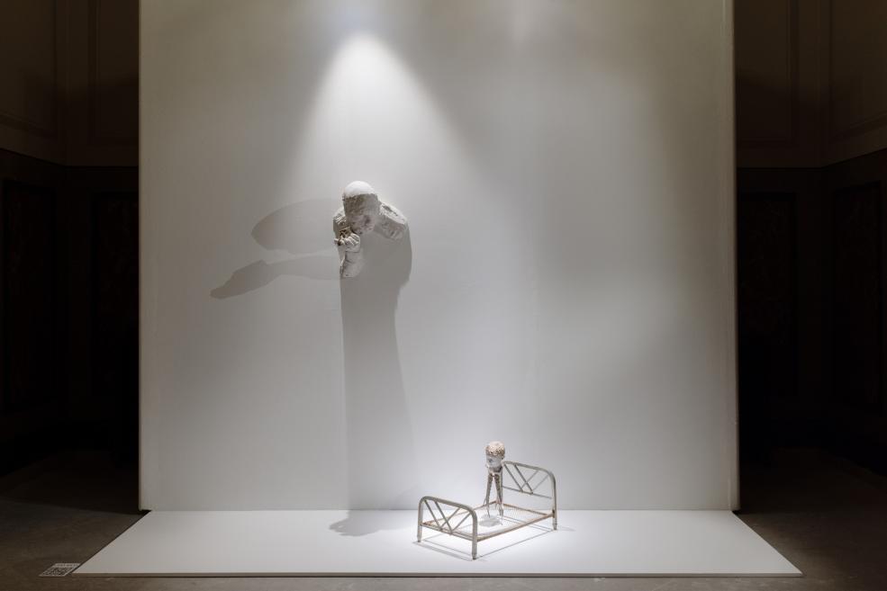 Museum installation with a white wall, a white head-and-arms sculpture lit by a spotlight, and a small metal bed frame with a figure placed on a white base below.
