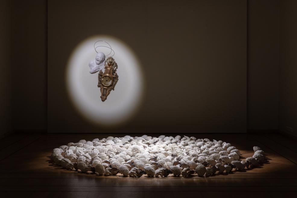 In a dark gallery, a decorative wall clock is lit on the wall beneath a white head sculpture, while a large white ring of ear-like objects lies on the floor.