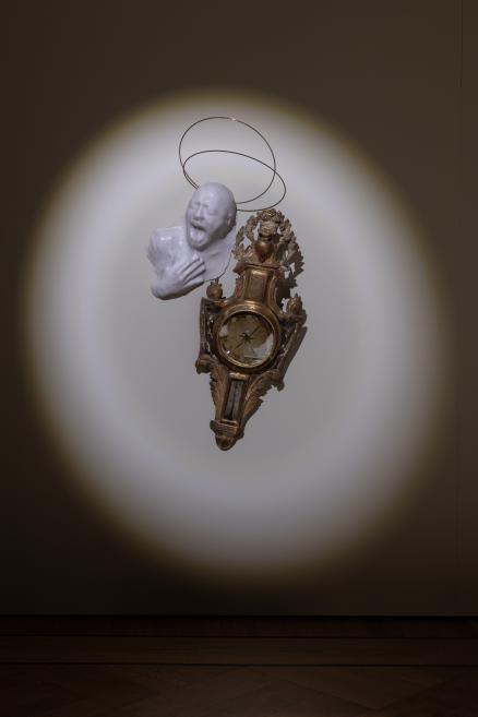 A decorative wall clock with an ornate gold-toned frame mounted on a wall, topped by a white sculptural human head.