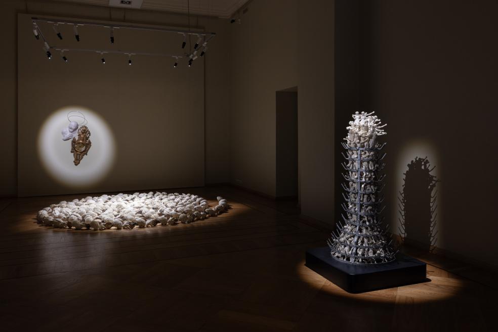 An installation in a museum room on a wooden floor features white reliefs, a small wall-mounted object with a head and clock motif in a spotlight, and a tall figurative sculpture on a pedestal at right.