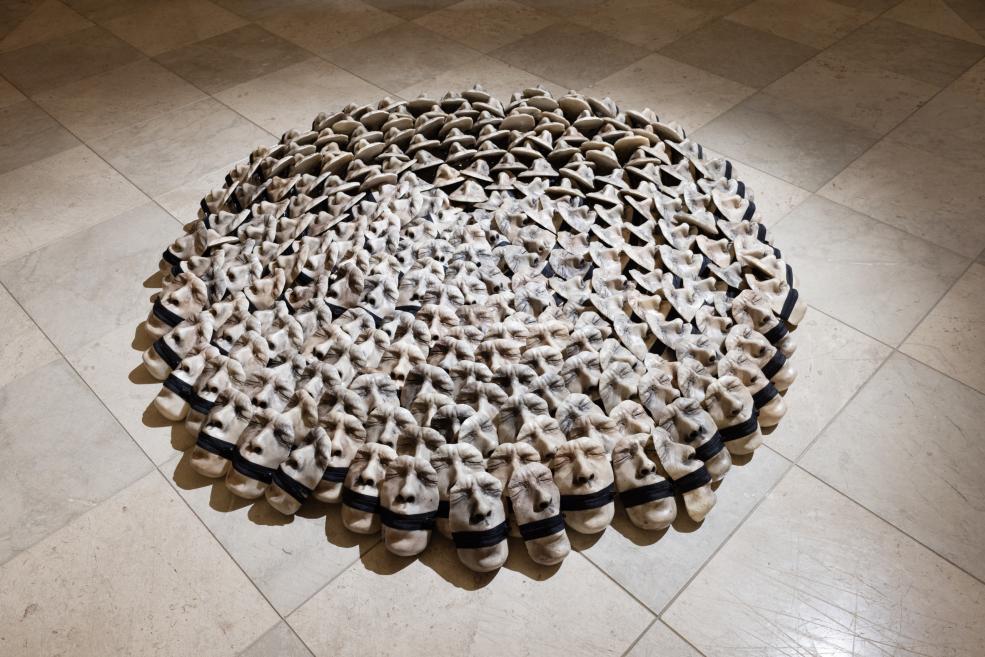 A large circular installation of many white mask-like objects with dark bands is arranged on the museum’s tiled floor.