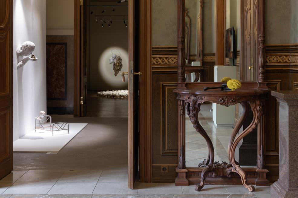 Museum interior with wood-paneled walls, an ornate console table with a yellow sculptural object, a mirror, and contemporary sculptures displayed in the adjoining room.