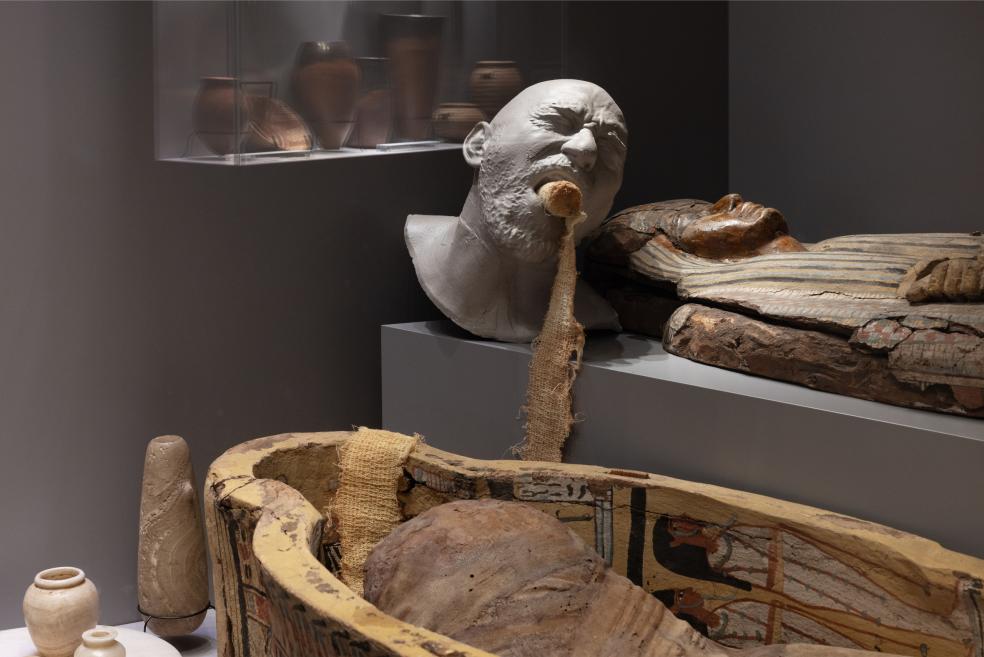 Several sculptural heads and sarcophagus fragments are displayed in a museum case, including a white plaster mask and polychrome wooden burial elements.