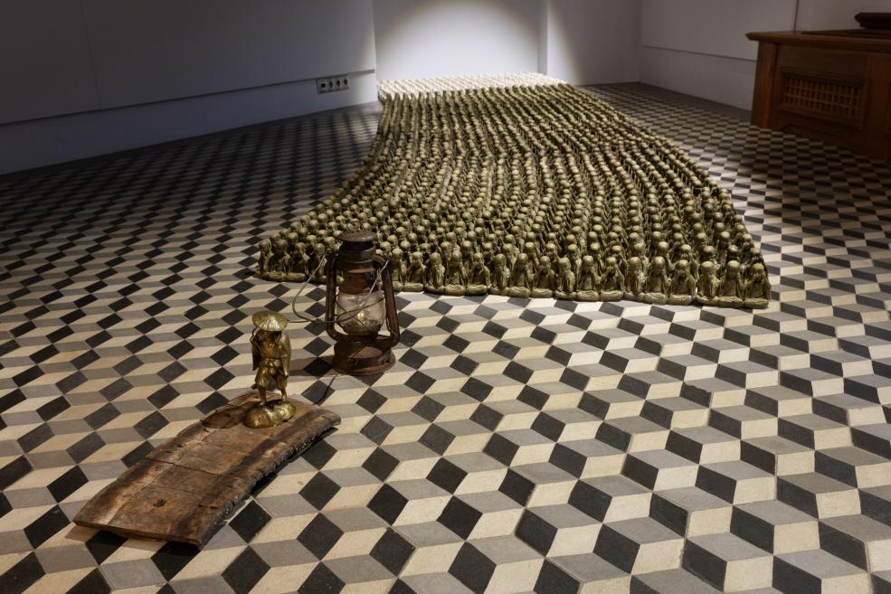 In an exhibition room with a geometric black-and-white floor, an installation of many small gold figurines arranged in rows is shown, with an old kerosene lamp and a small bronze figure on a wooden plank in the foreground.