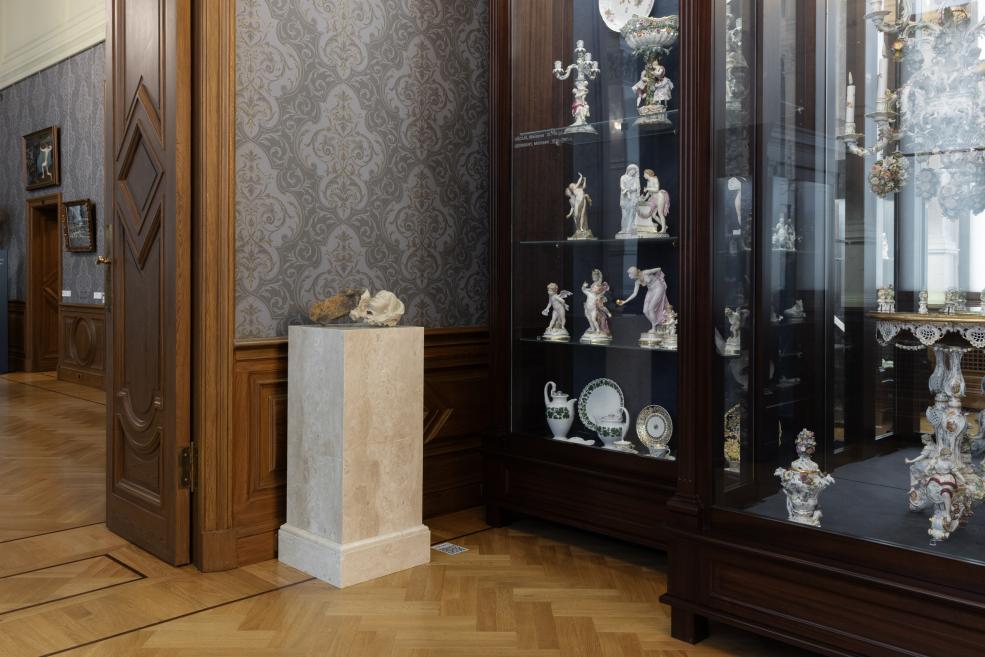 Museum room with parquet flooring, patterned walls, a marble pedestal with a sculptural object, and a glass display case containing porcelain figurines and vessels.