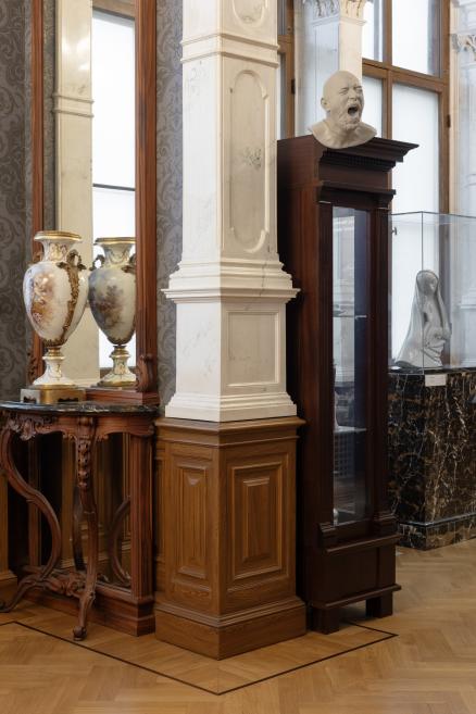 Decorative vases, a sculptural bust on a dark pedestal, and a white sculpture in a glass display case are shown in a museum interior.