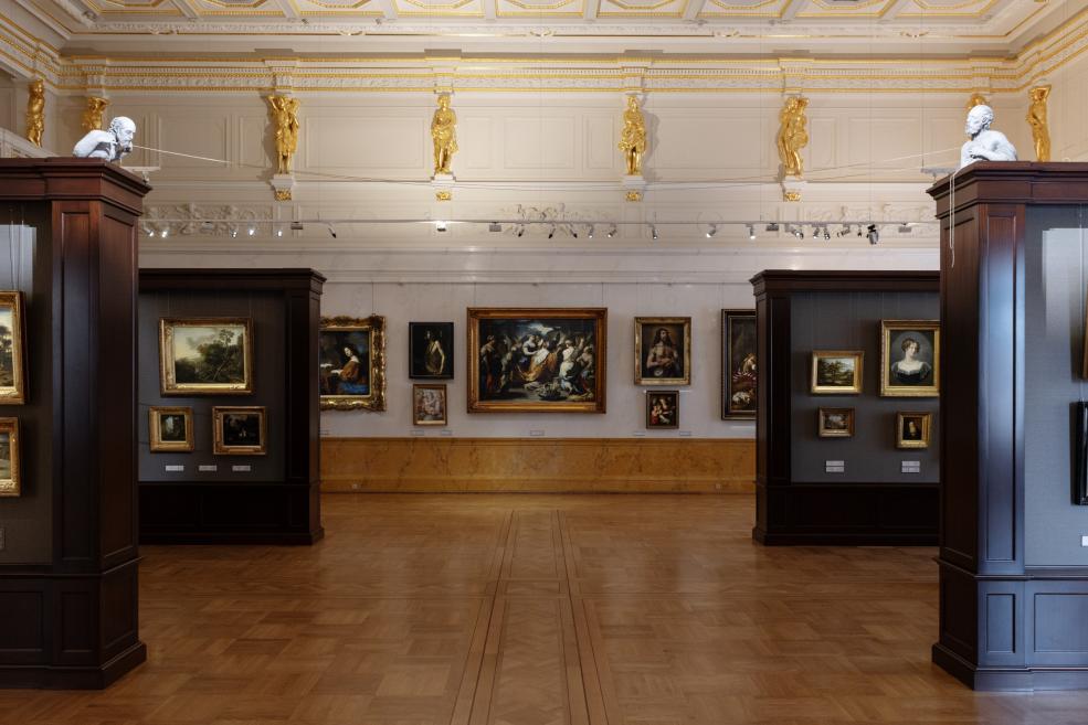 Museum gallery with a neoclassical interior, gilded wall sculptures, and large framed paintings on both sides.
