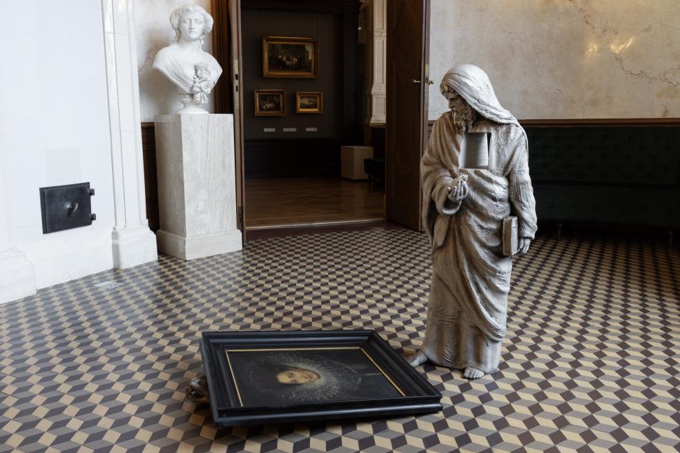 In a museum hall with a patterned floor, a draped sculpture holding a book stands beside an overturned framed portrait, with a white bust on a pedestal in the background.