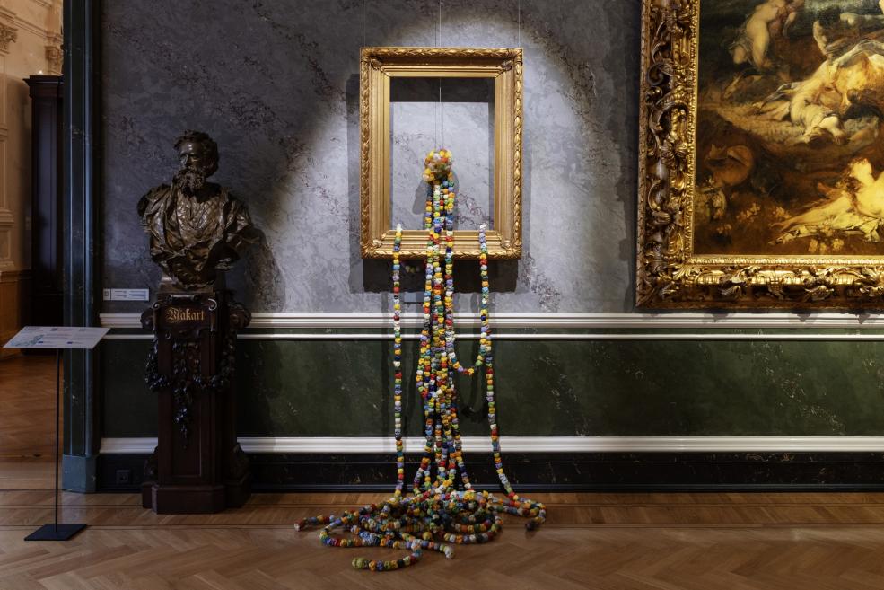 In a museum gallery, an empty gilded frame on the wall has a multicolored bead sculpture hanging down from it, beside a bronze bust and a painting in a gilded frame.