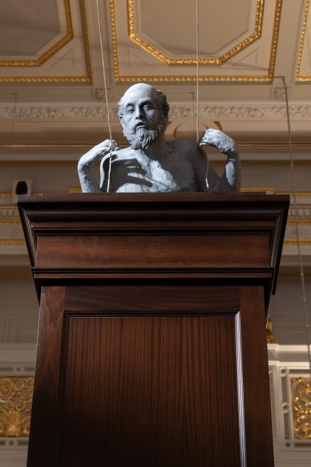 White-painted bust sculpture of a bearded man on a dark wooden pedestal in a museum hall with gilded ceiling details.
