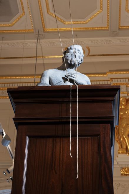 A sculptural male bust on a dark wooden pedestal in a museum hall with decorative gold ceiling and wall details, with a white cord hanging down the front.