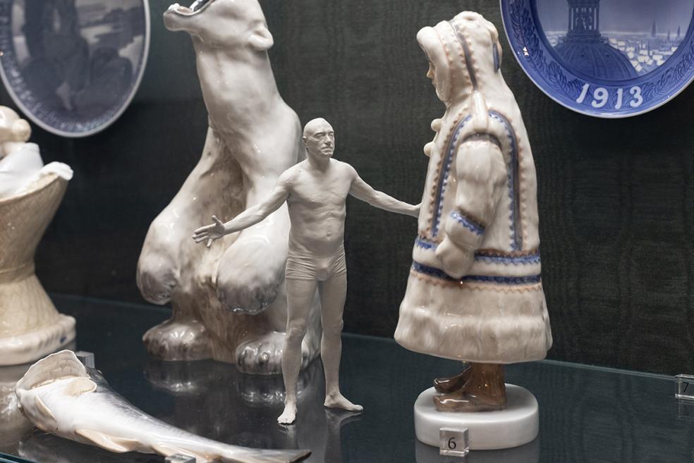 Display case of porcelain figurines with a nude male figure between larger animal and human figures against a dark background.