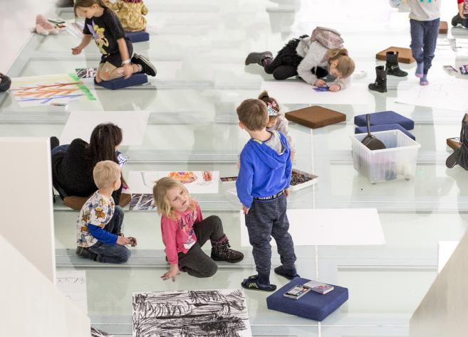 Bērni sēž un zīmē uz stiklotas grīdas muzeja radošajā nodarbībā, apkārt izklātas lapas un krītiņi.